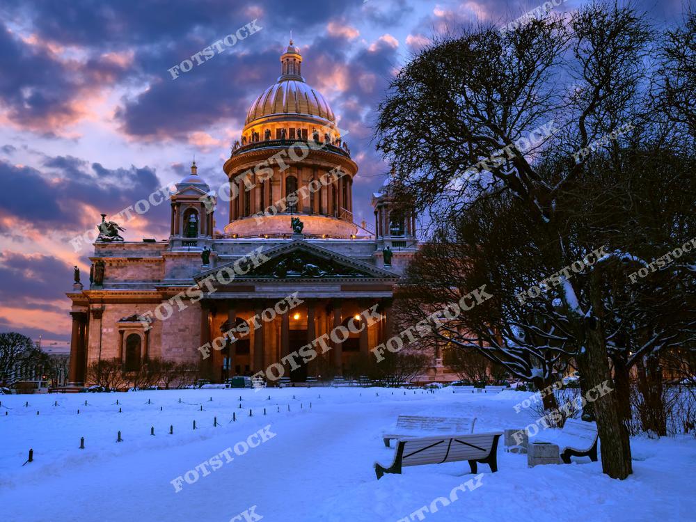 На фотографии изображен Исаакиевский собор в лучах закатного солнца. Яркое небо с облаками создает контраст с подсвеченным собором, который выделяется на фоне снежной площади. Здание собора величественно возвышается, а заснеженные деревья и скамейки подчеркивают зимнюю атмосферу Санкт-Петербурга. Это одно из самых известных архитектурных сооружений города, особенно красивое в вечерние часы.